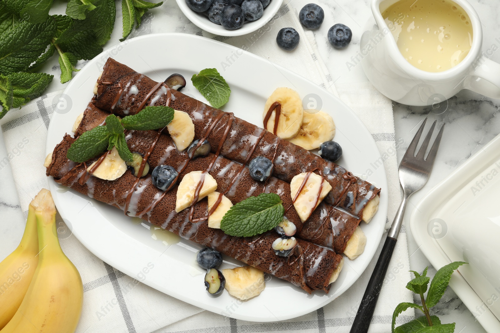 Photo of Delicious chocolate crepes with banana and blueberries served on white marble table, flat lay