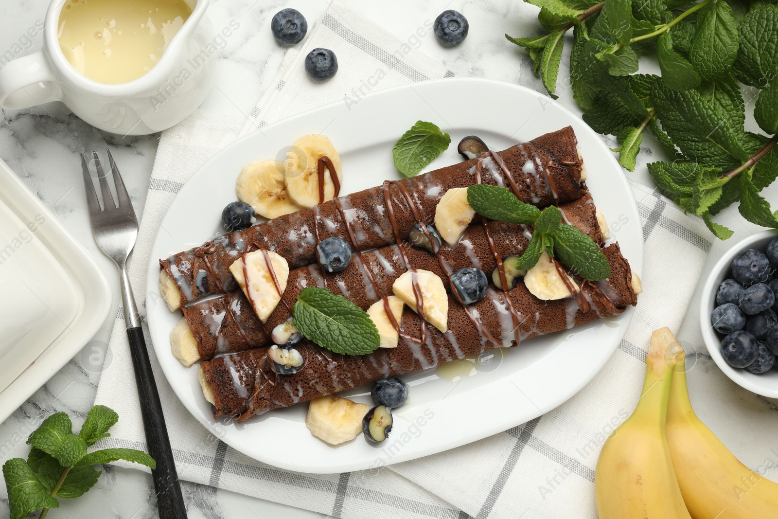 Photo of Delicious chocolate crepes with banana and blueberries served on white marble table, flat lay