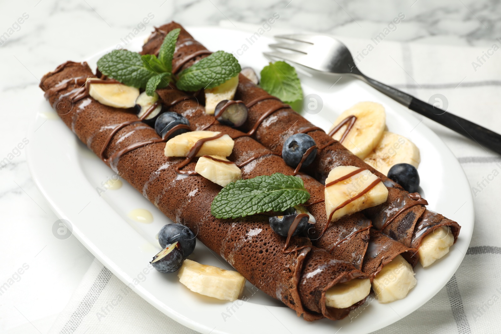 Photo of Delicious chocolate crepes with banana and blueberries served on white marble table, closeup