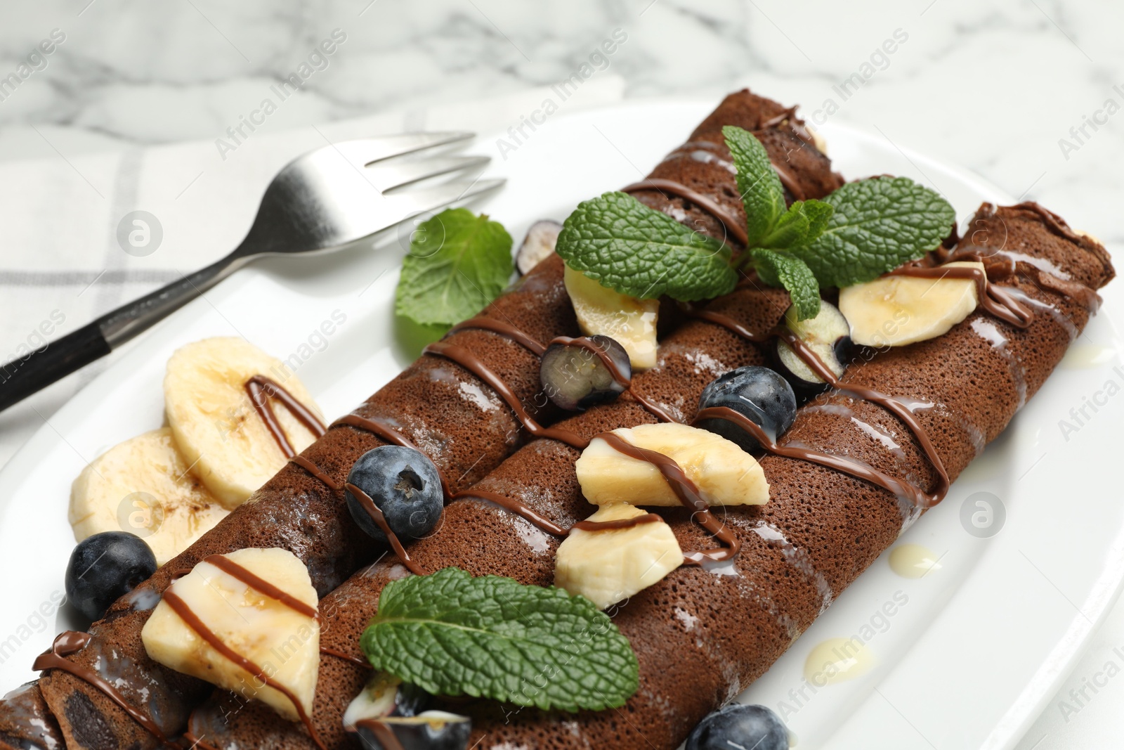 Photo of Delicious chocolate crepes with banana and blueberries served on white marble table, closeup