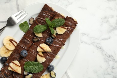 Photo of Delicious chocolate crepes with banana and blueberries served on white marble table, top view. Space for text