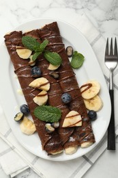 Photo of Delicious chocolate crepes with banana and blueberries served on white marble table, top view