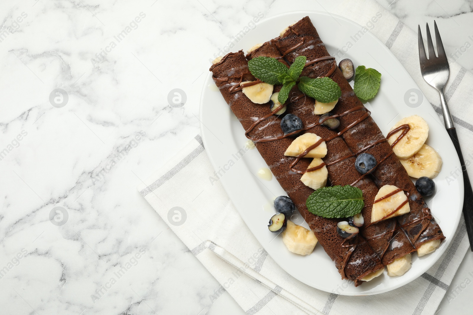 Photo of Delicious chocolate crepes with banana and blueberries served on white marble table, top view. Space for text