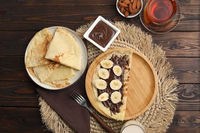 Photo of Delicious crepes with chocolate paste and banana served on wooden table, flat lay