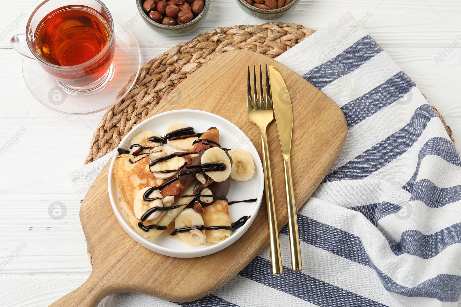 Photo of Delicious crepes with banana, nuts, chocolate sauce and tea on white wooden table, flat lay