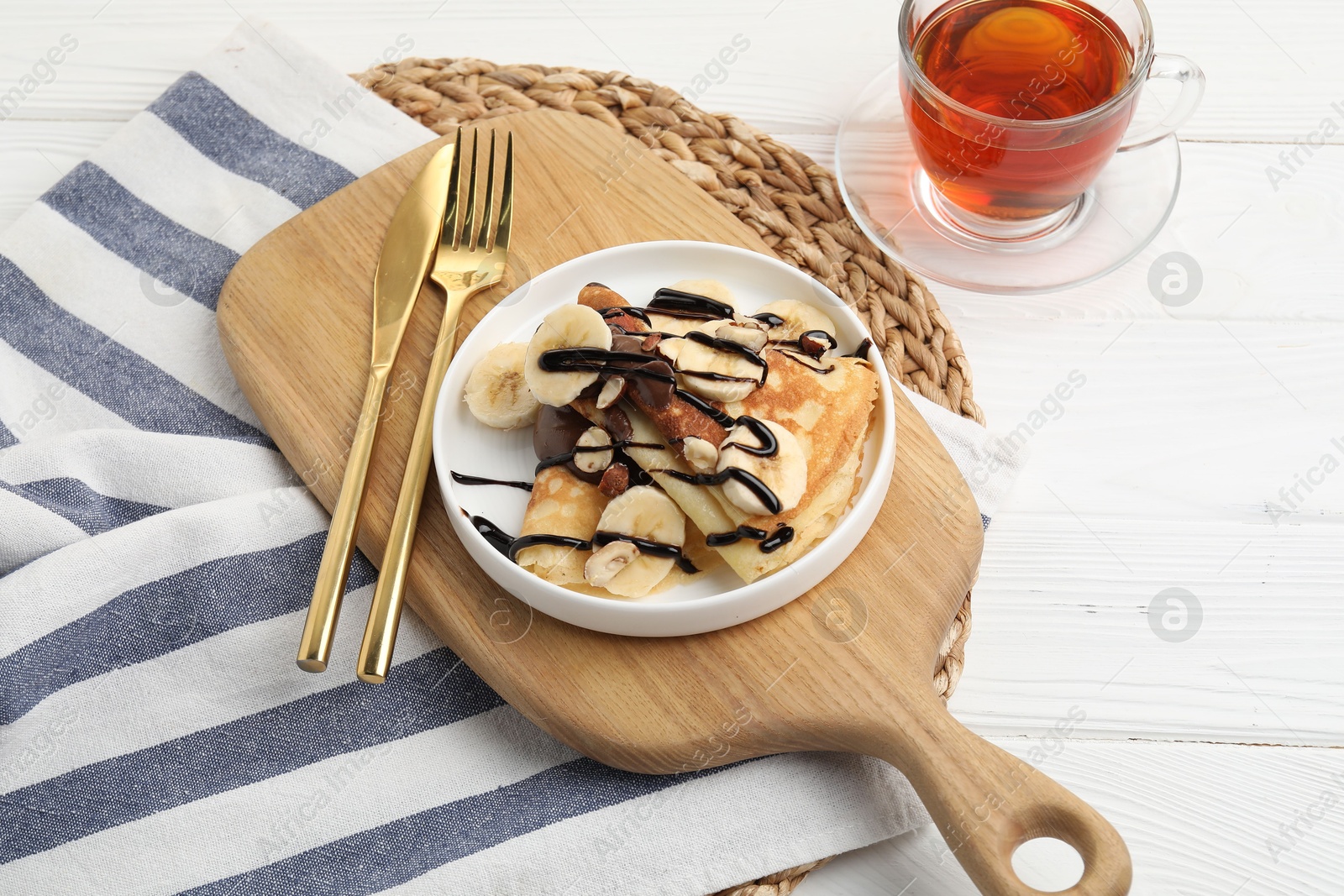 Photo of Delicious crepes with banana, nuts, chocolate sauce and tea on white wooden table