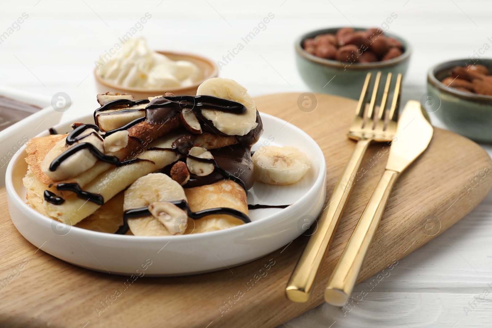 Photo of Delicious crepes with banana, nuts, chocolate sauce and whipped cream on white wooden table, closeup