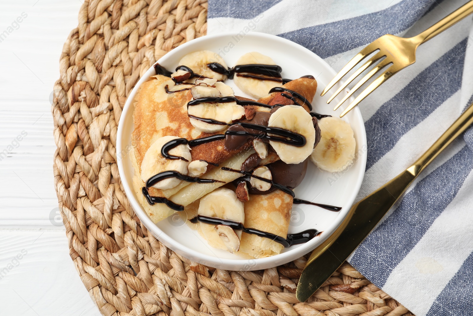 Photo of Delicious crepes with banana, chocolate sauce and nuts on white wooden table, top view