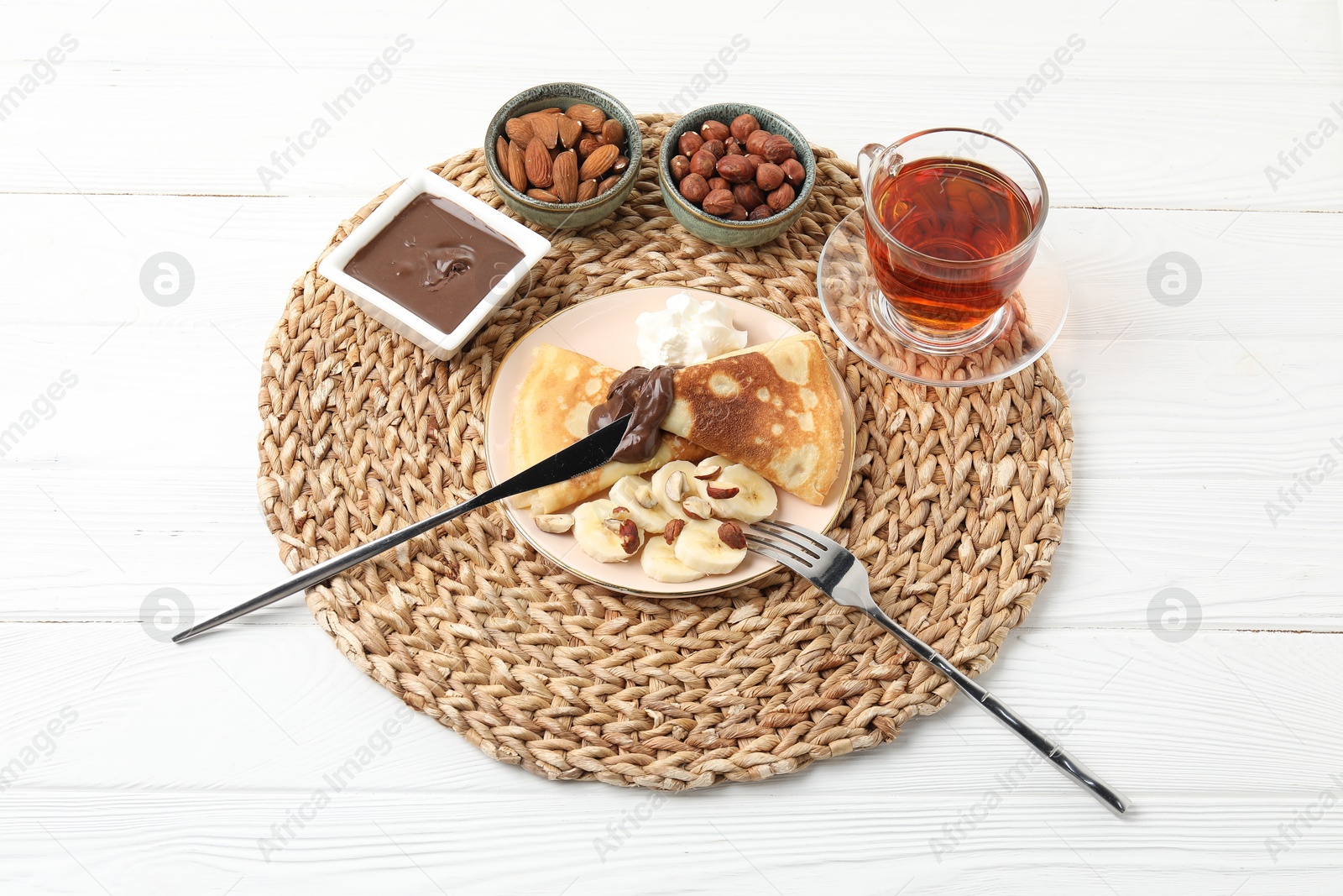 Photo of Delicious crepes with banana, nuts, chocolate paste, whipped cream and tea on white wooden table