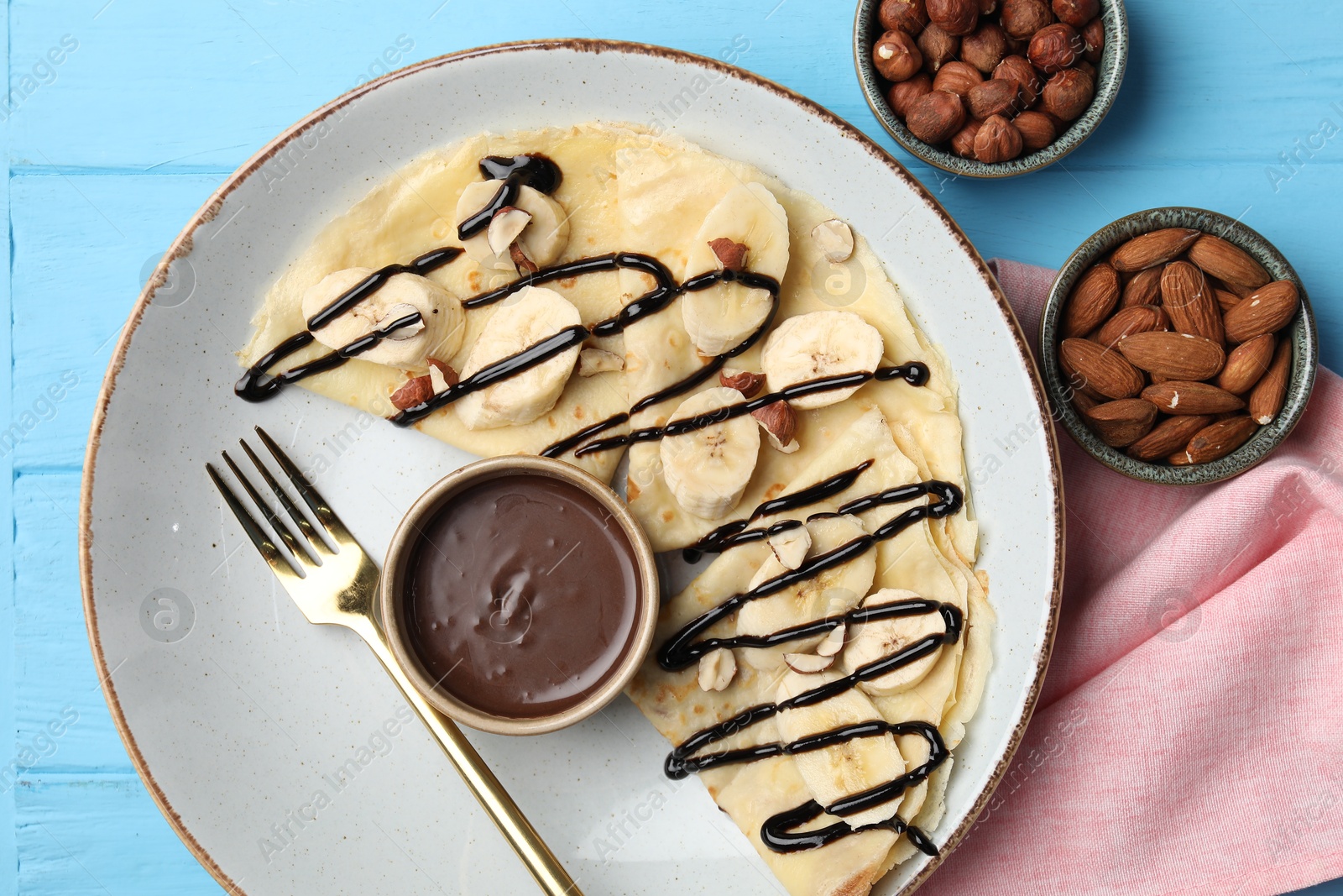 Photo of Delicious crepes with banana, nuts and chocolate sauce on light blue wooden table, flat lay