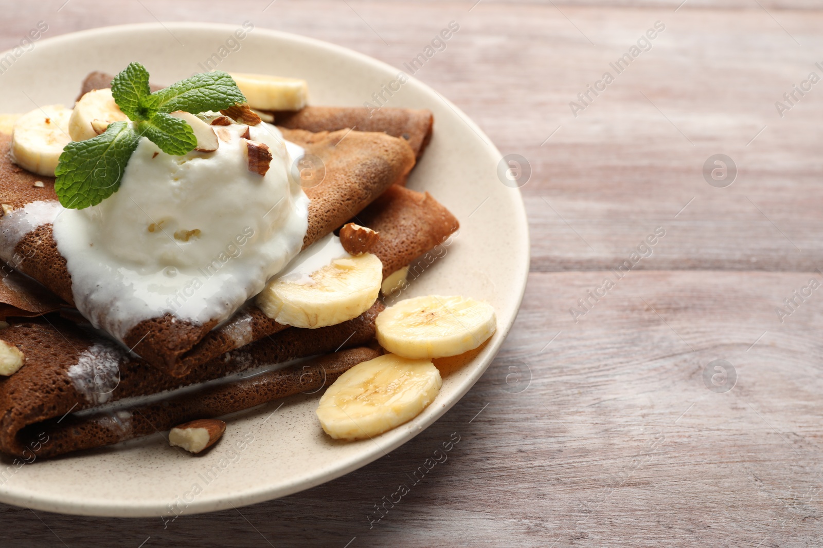 Photo of Delicious chocolate crepes with banana, walnuts, mint and scoop of ice cream on wooden table, closeup. Space for text