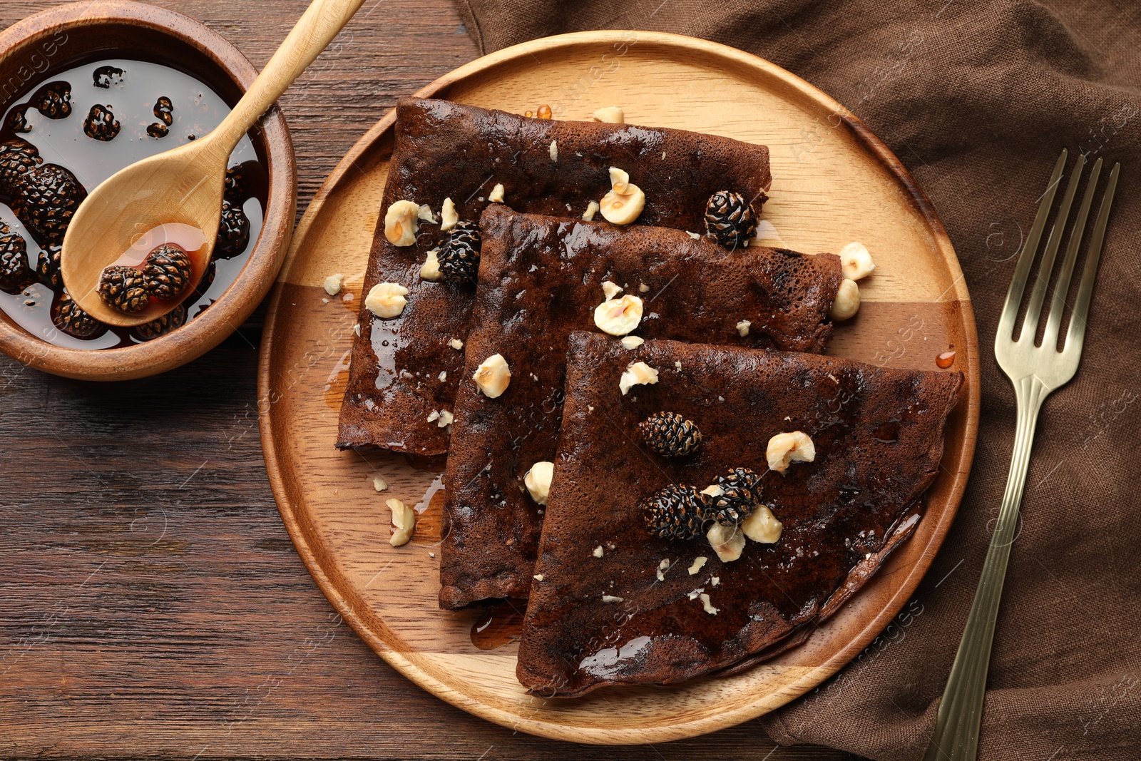 Photo of Delicious chocolate crepes with pine cone jam and nuts on wooden table, flat lay