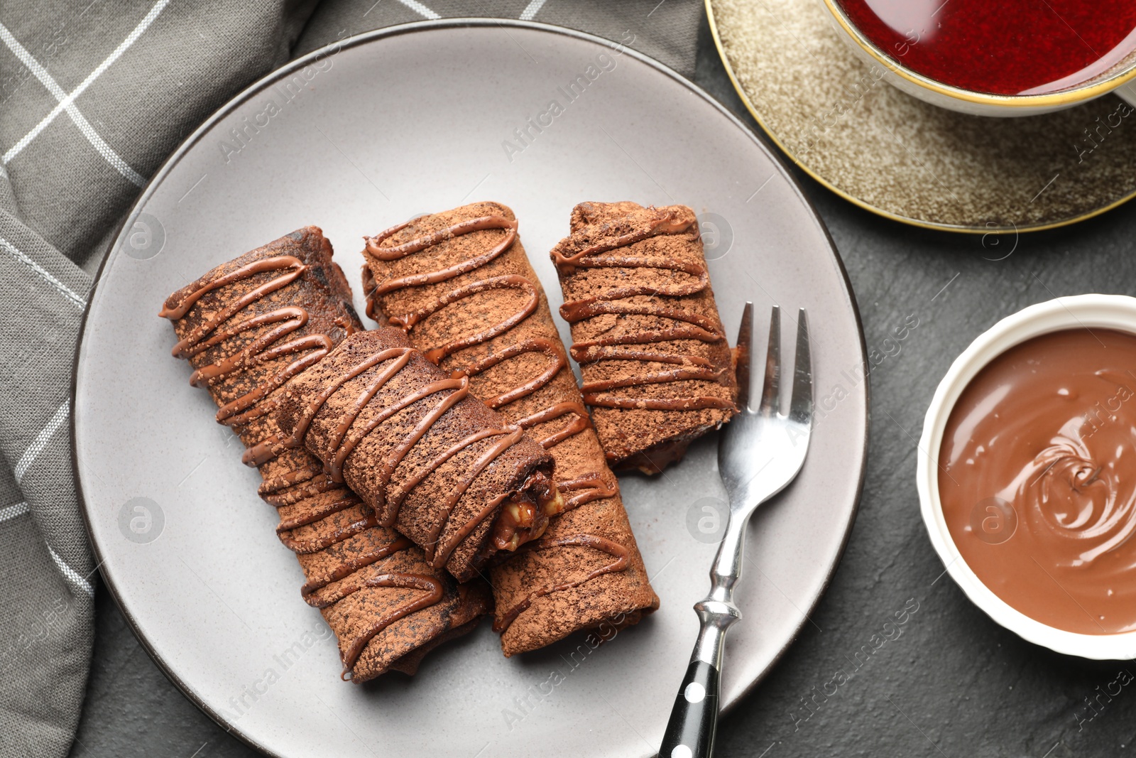 Photo of Delicious chocolate crepes with sweet paste, nuts and tea on black table, flat lay