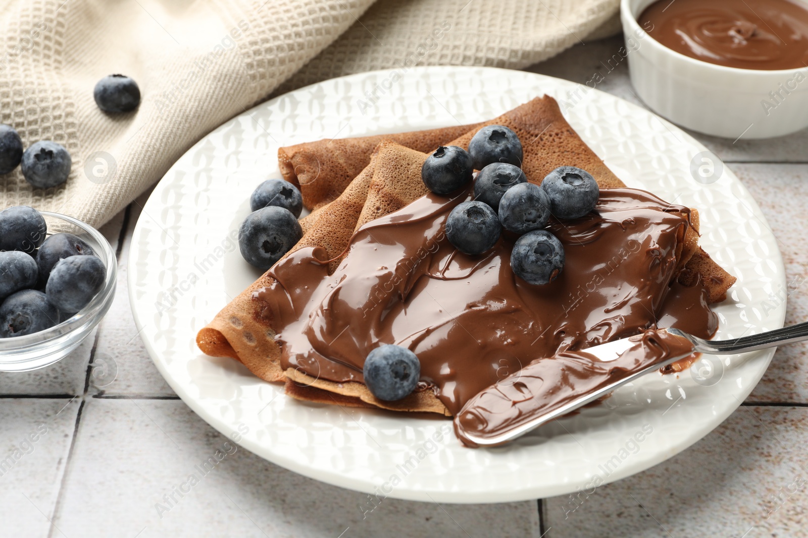 Photo of Delicious chocolate crepes with sweet paste and blueberries on light tiled table, closeup