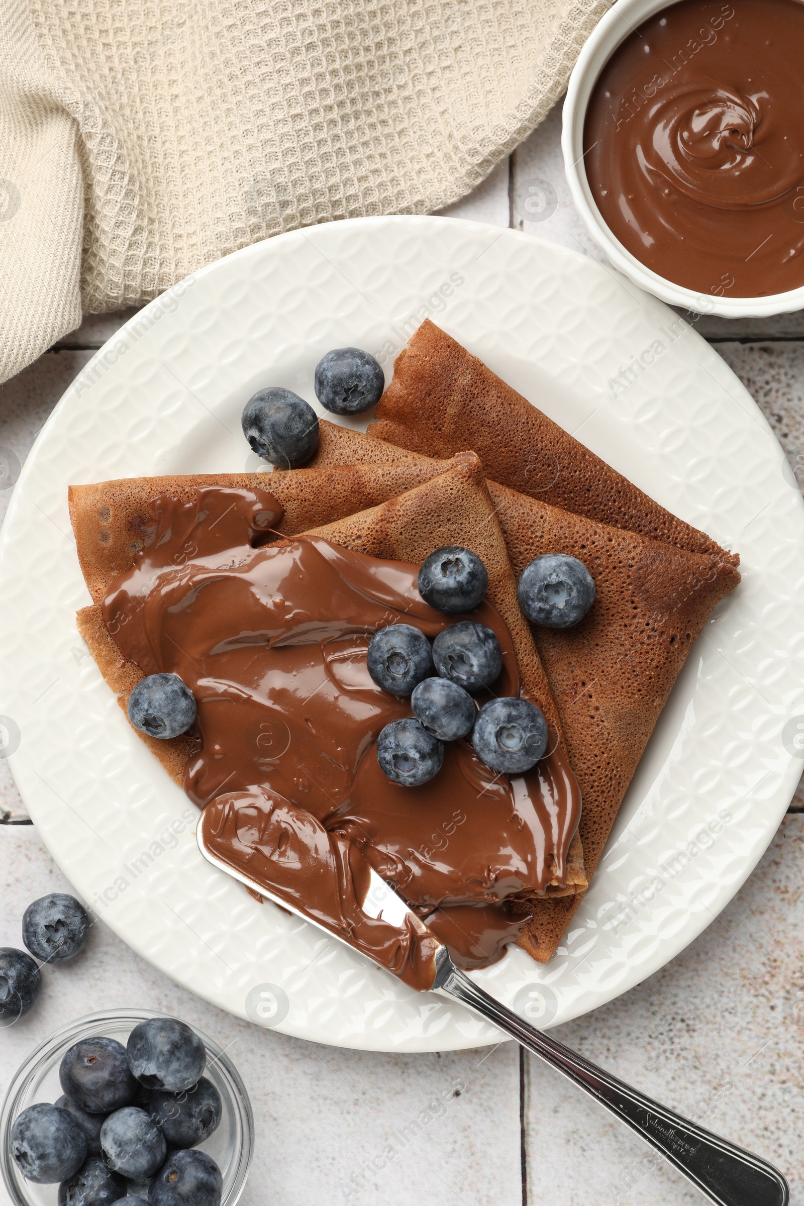 Photo of Delicious chocolate crepes with sweet paste and blueberries on light tiled table, flat lay