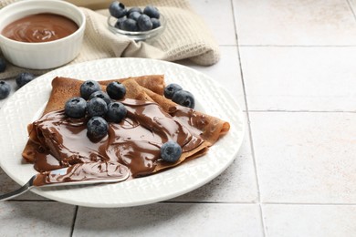 Photo of Delicious chocolate crepes with sweet paste and blueberries on light tiled table, closeup. Space for text