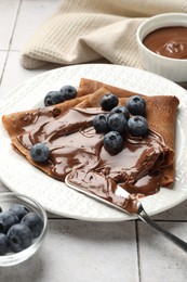 Photo of Delicious chocolate crepes with sweet paste and blueberries on light tiled table, closeup