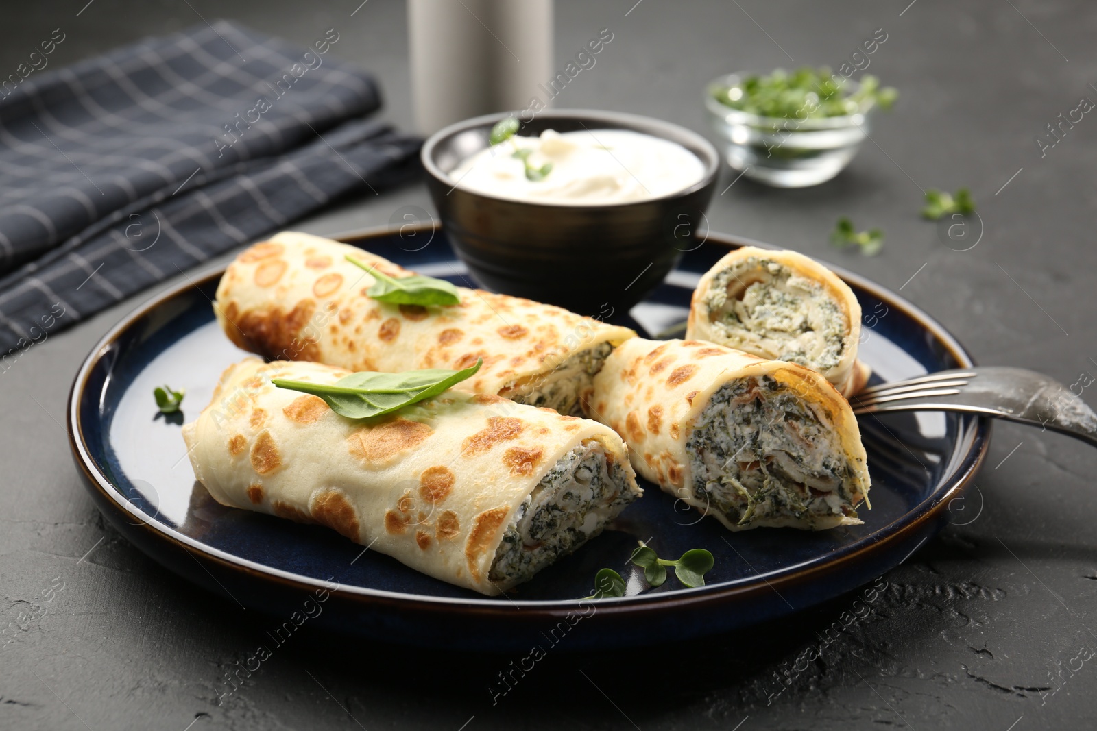 Photo of Delicious crepes with cottage cheese, spinach and sour cream served on black table, closeup