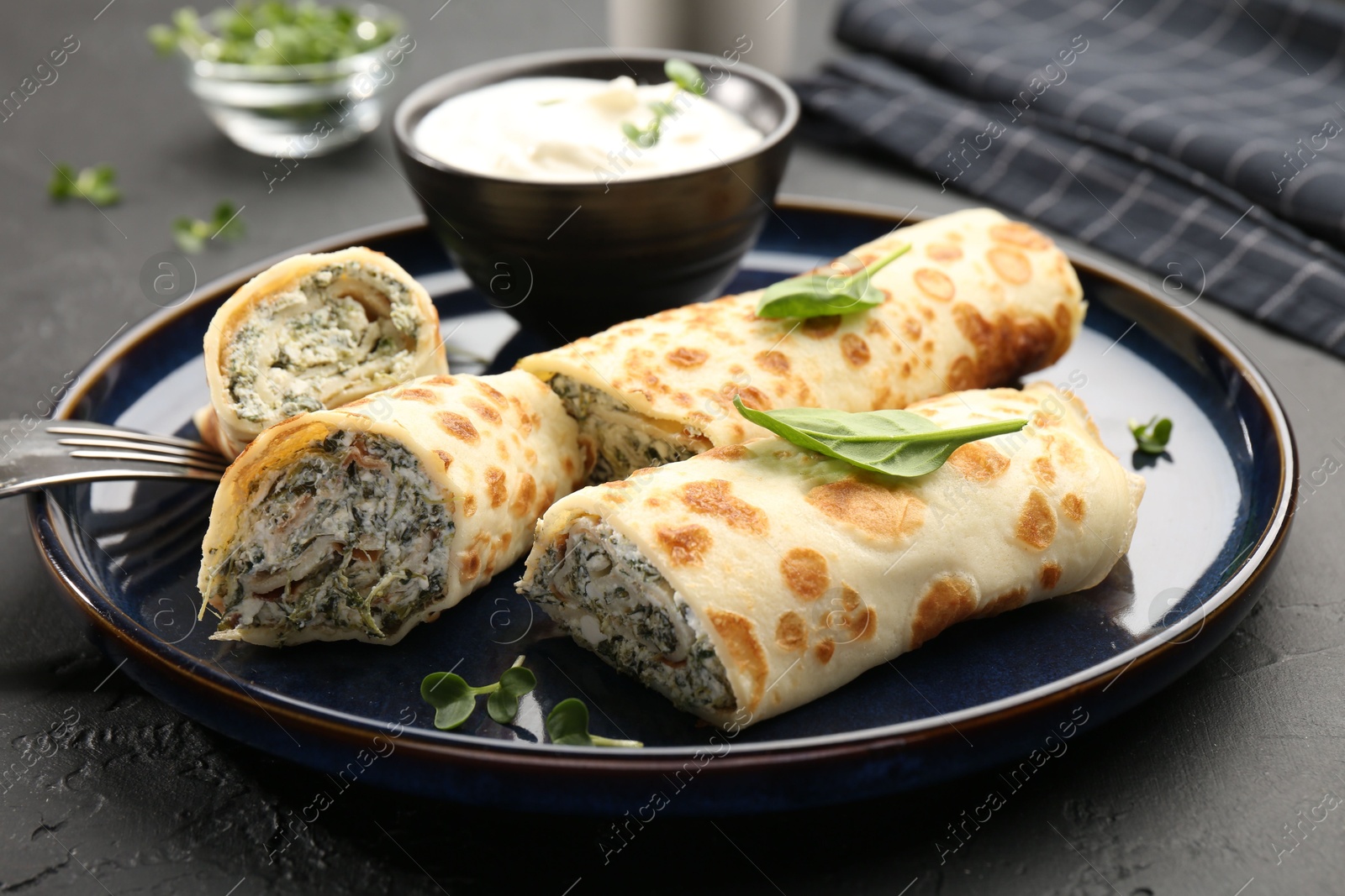 Photo of Delicious crepes with cottage cheese, spinach and sour cream served on black table, closeup