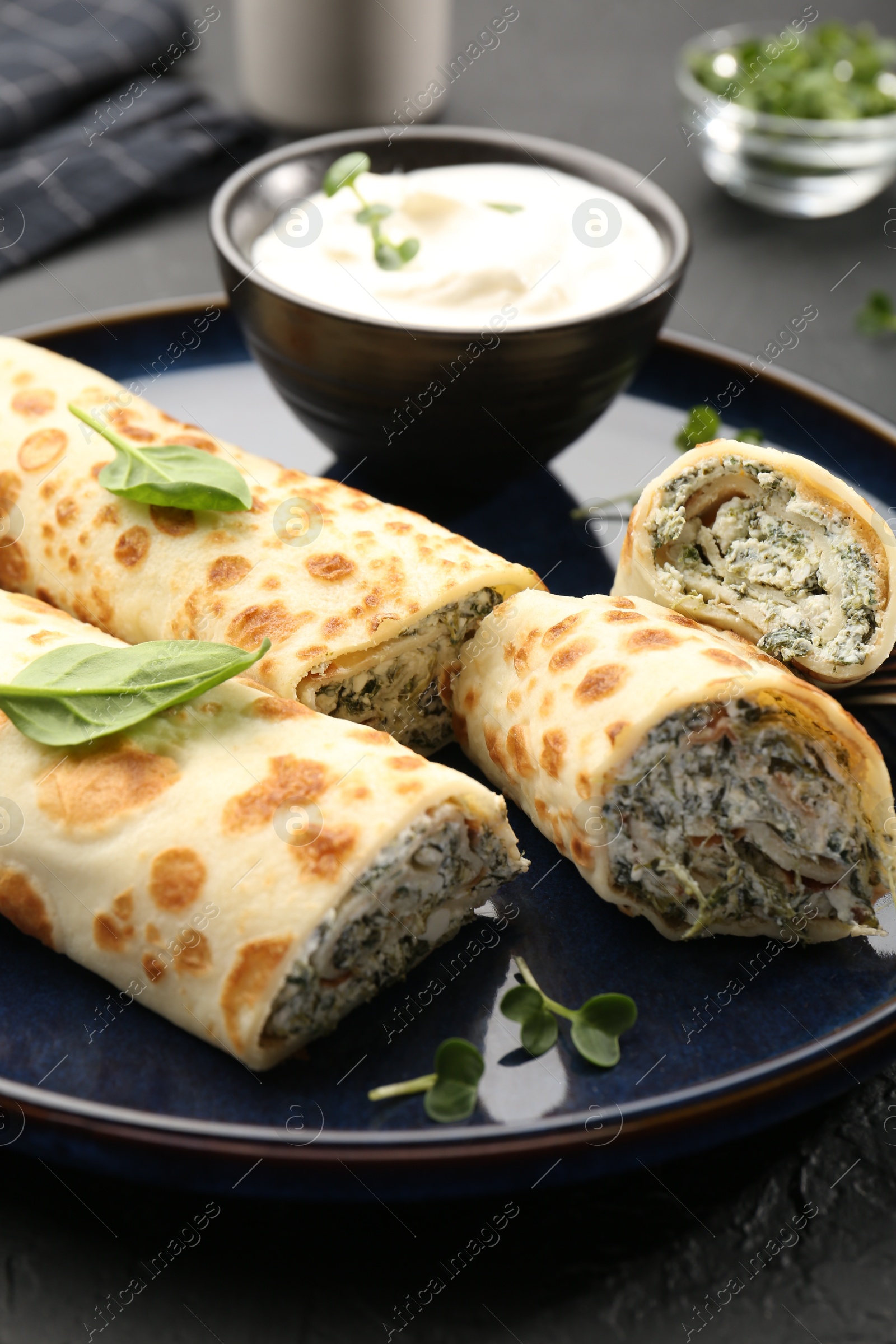 Photo of Delicious crepes with cottage cheese, spinach and sour cream on black table, closeup