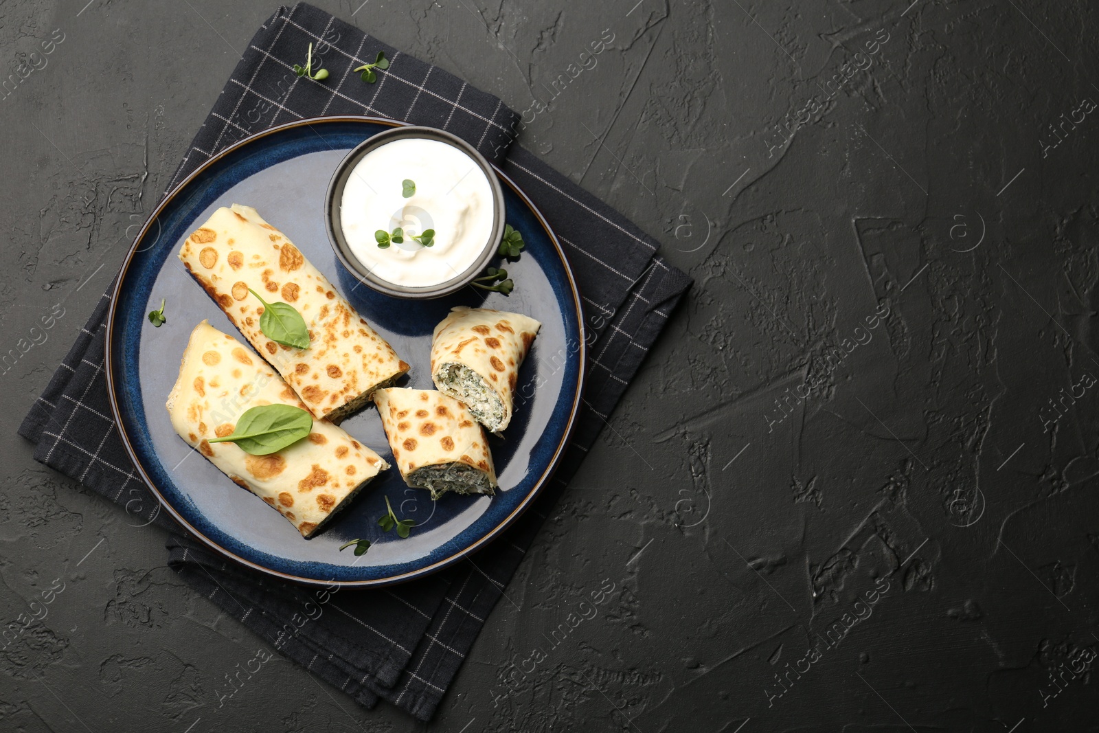 Photo of Delicious crepes with cottage cheese, spinach and sour cream on black table, top view. Space for text