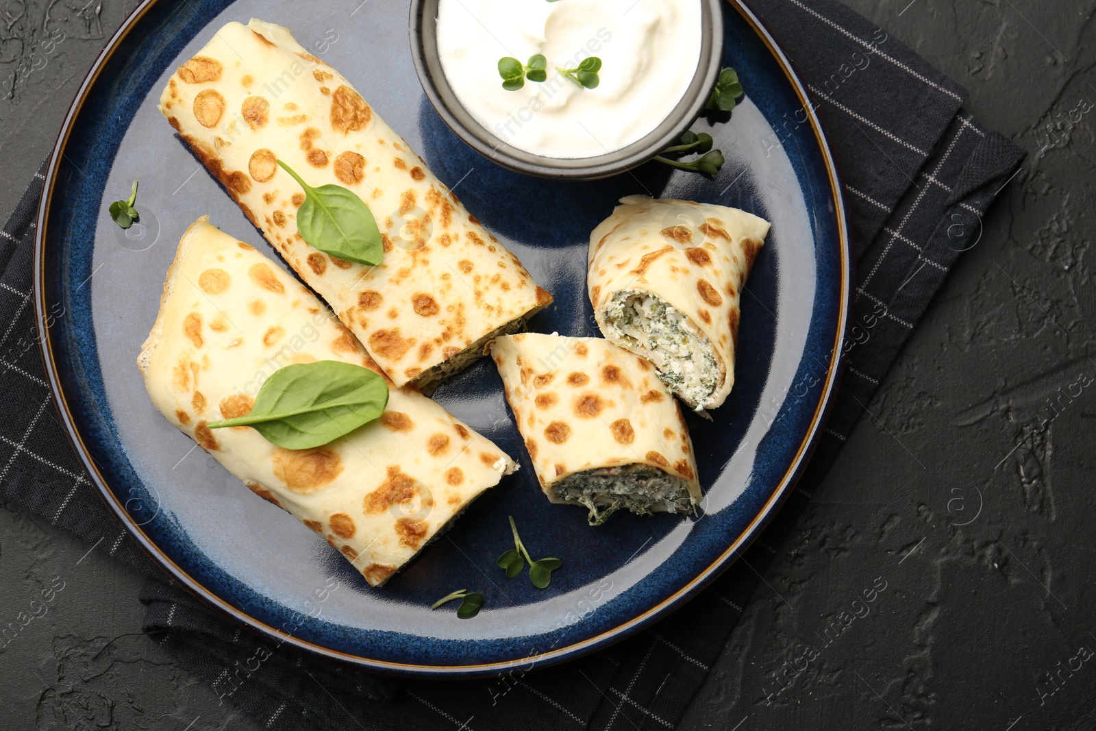 Photo of Delicious crepes with cottage cheese, spinach and sour cream on black table, top view