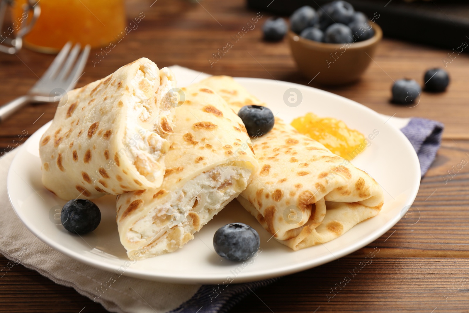 Photo of Delicious crepes with cottage cheese, jam and blueberries on wooden table, closeup