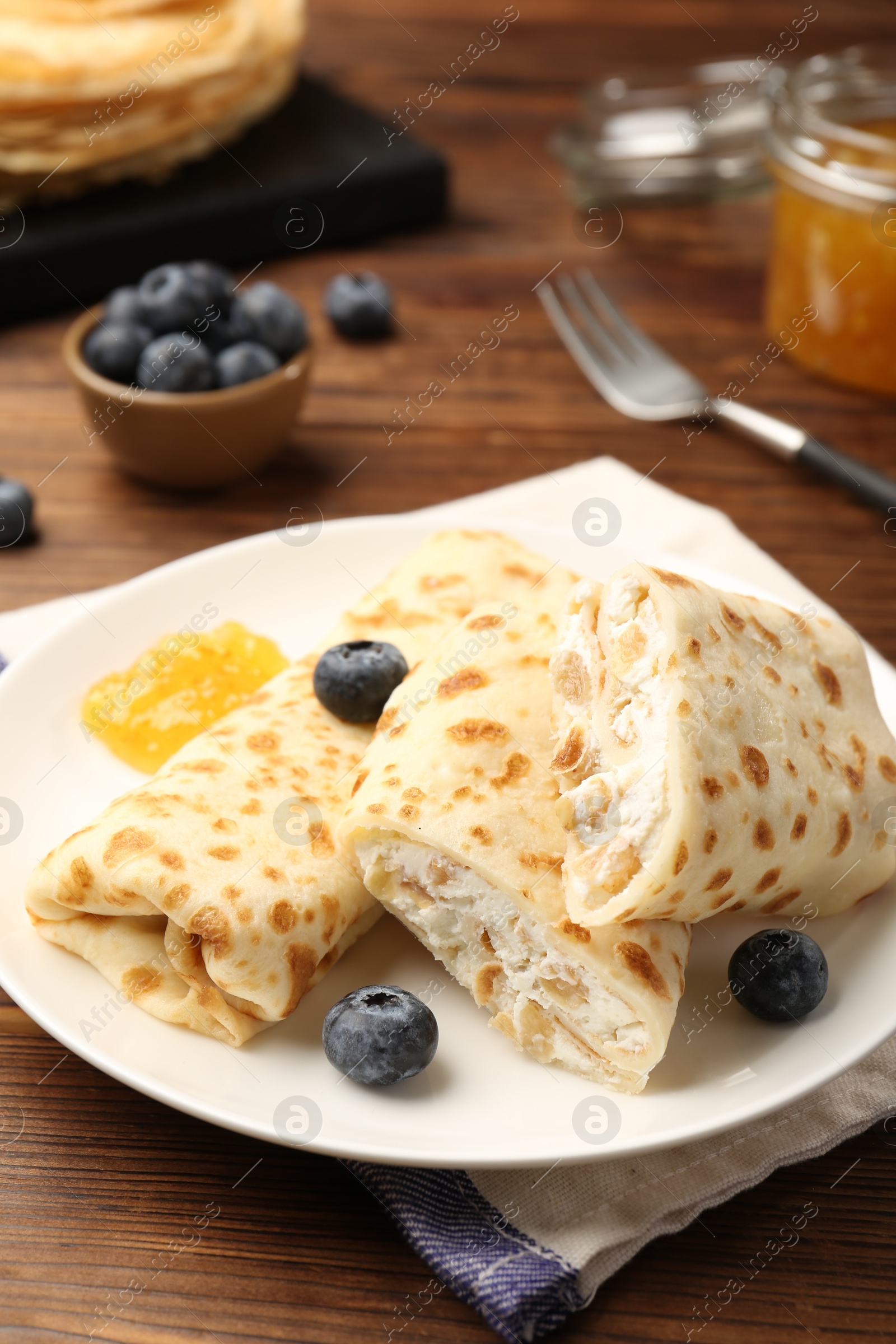 Photo of Delicious crepes with cottage cheese, jam and blueberries served on wooden table, closeup