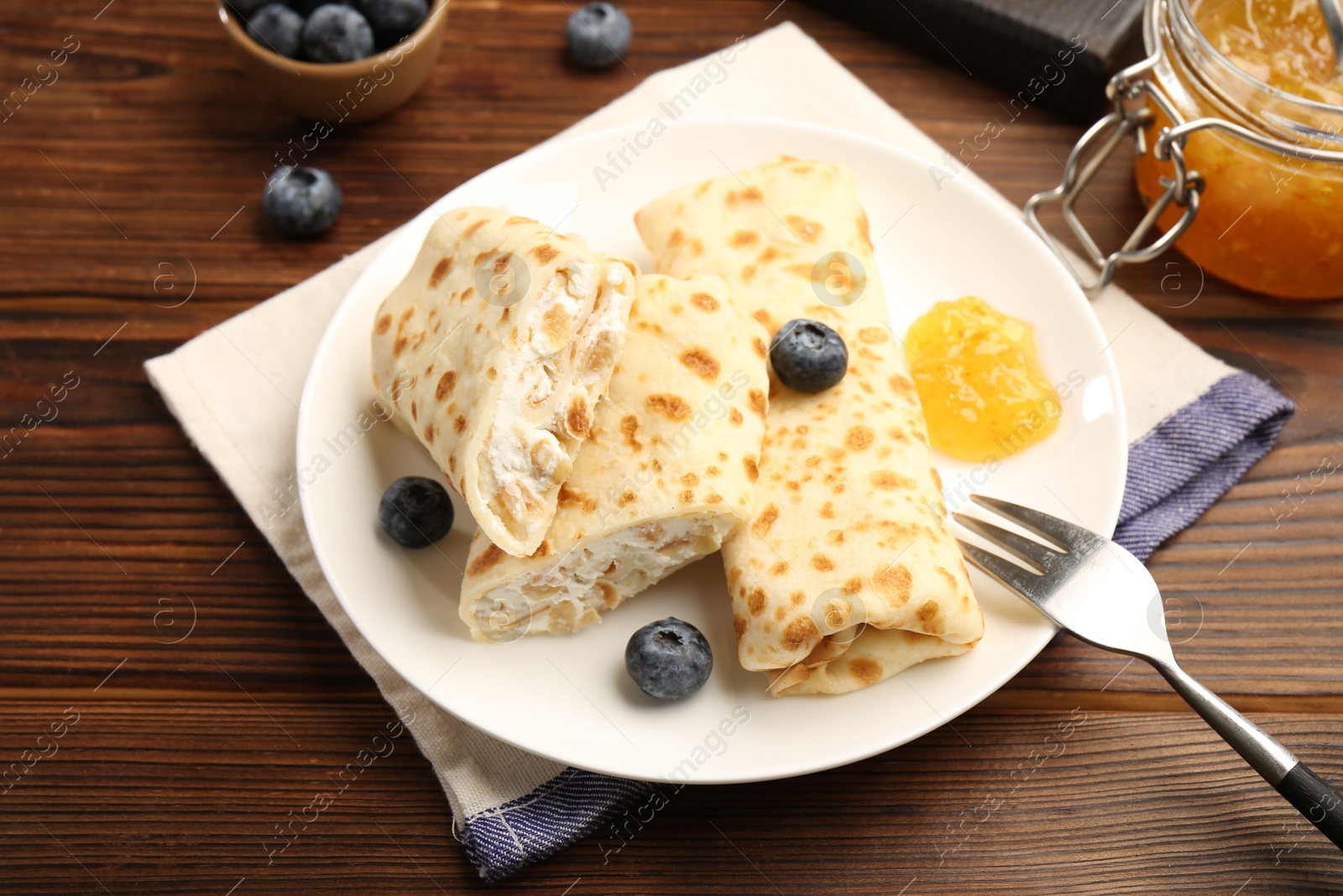 Photo of Delicious crepes with cottage cheese, jam and blueberries served on wooden table, closeup