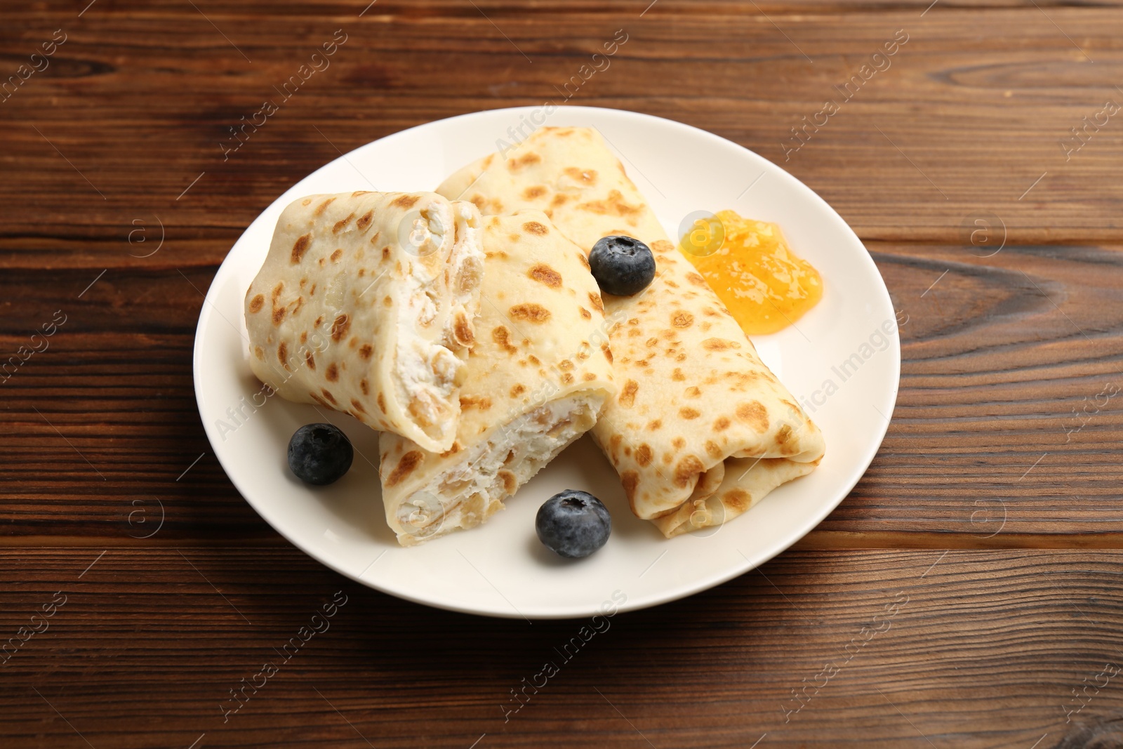 Photo of Delicious crepes with cottage cheese, jam and blueberries on wooden table, closeup