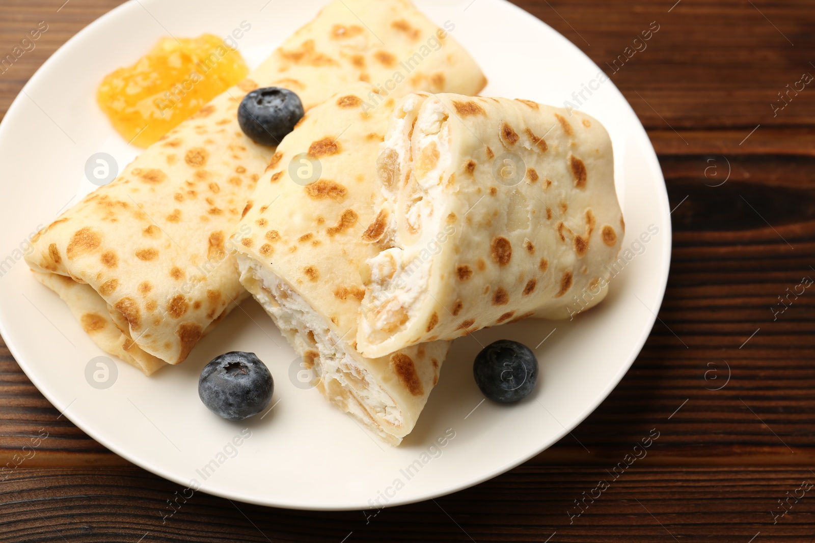 Photo of Delicious crepes with cottage cheese, jam and blueberries on wooden table, closeup