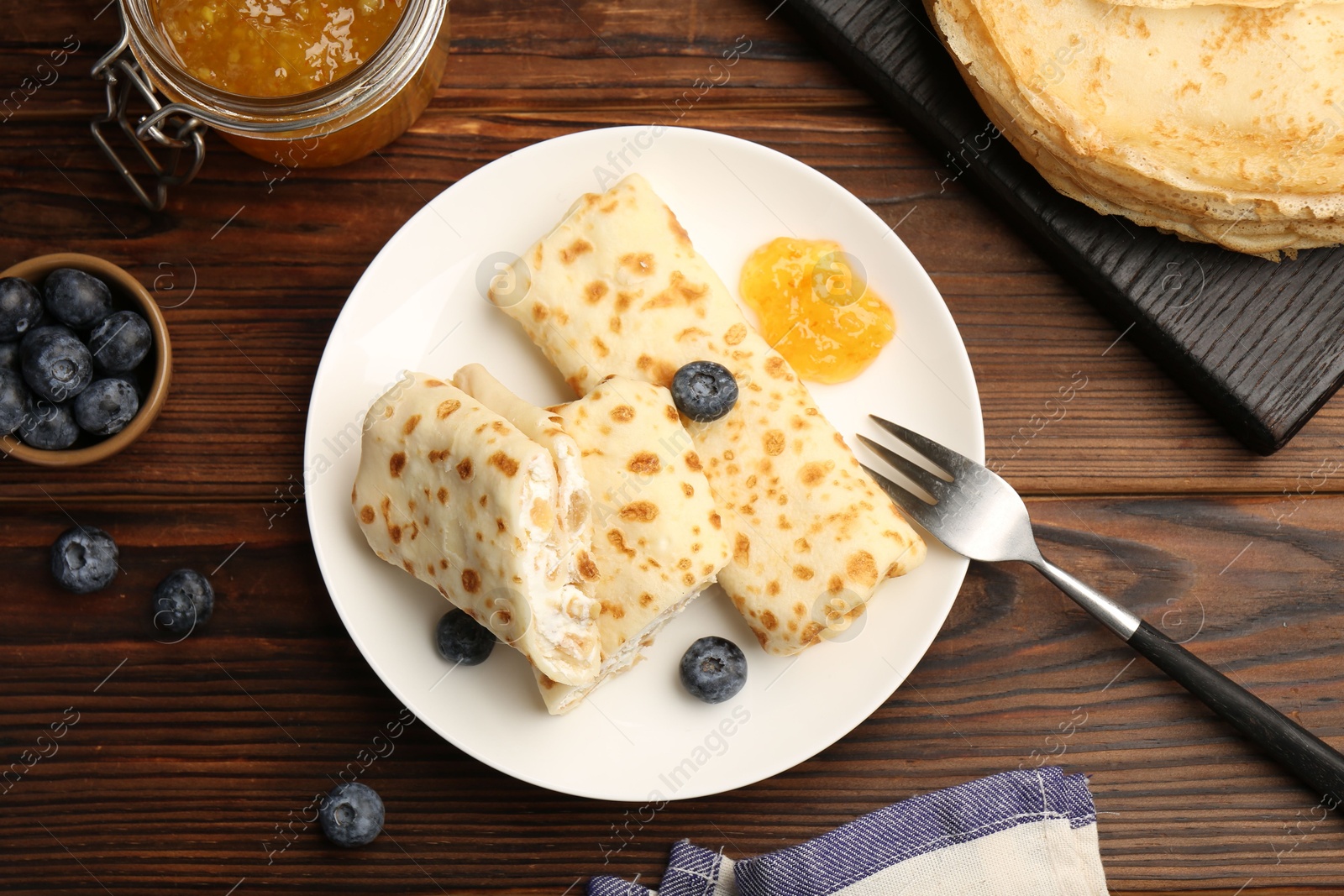 Photo of Delicious crepes with cottage cheese, jam and blueberries served on wooden table, flat lay