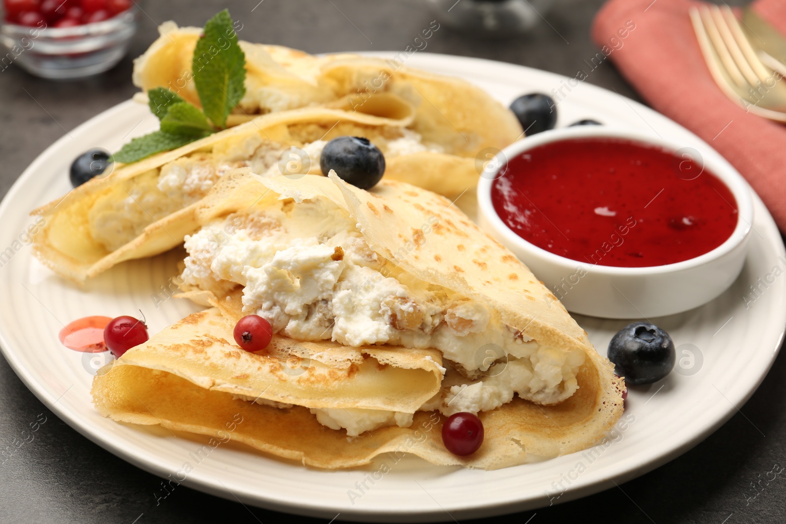 Photo of Delicious crepes with cottage cheese, jam, redcurrants and blueberries on grey table, closeup