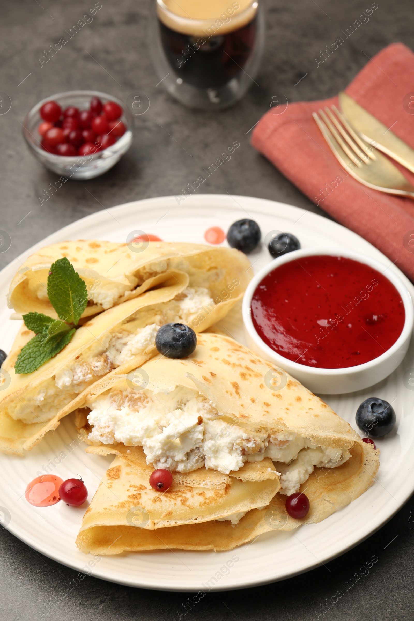 Photo of Delicious crepes with cottage cheese, jam, redcurrants and blueberries served on grey table, closeup