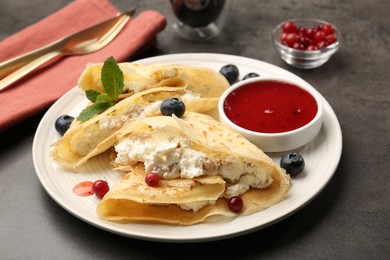 Photo of Delicious crepes with cottage cheese, jam, redcurrants and blueberries served on grey table, closeup