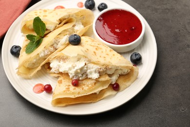 Photo of Delicious crepes with cottage cheese, jam, redcurrants and blueberries on grey table, closeup