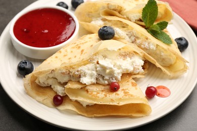 Photo of Delicious crepes with cottage cheese, jam, redcurrants and blueberries on grey table, closeup