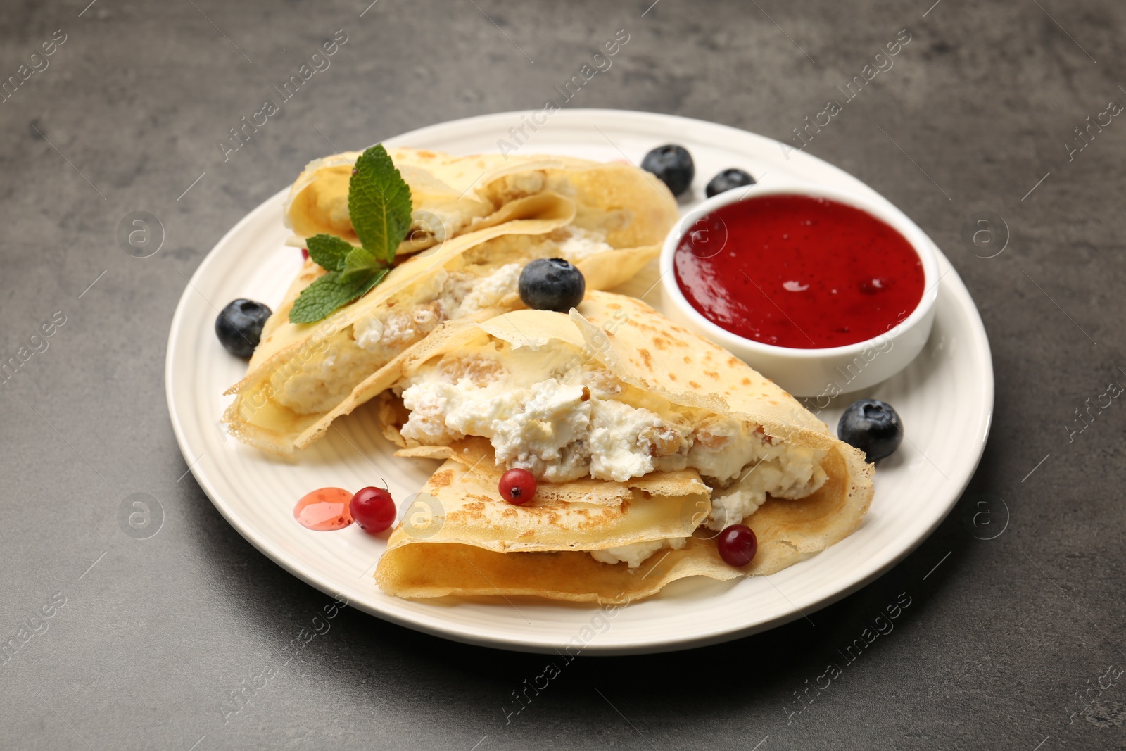 Photo of Delicious crepes with cottage cheese, jam, redcurrants and blueberries on grey table, closeup