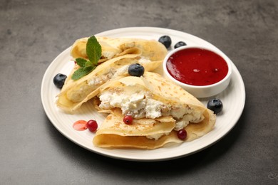 Photo of Delicious crepes with cottage cheese, jam, redcurrants and blueberries on grey table, closeup