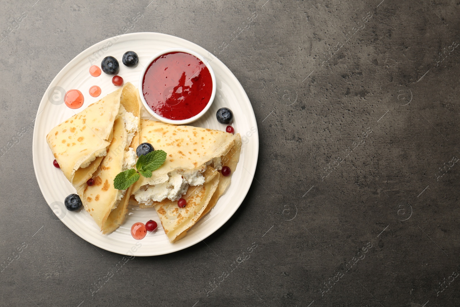 Photo of Delicious crepes with cottage cheese, jam, redcurrants and blueberries on grey table, top view. Space for text