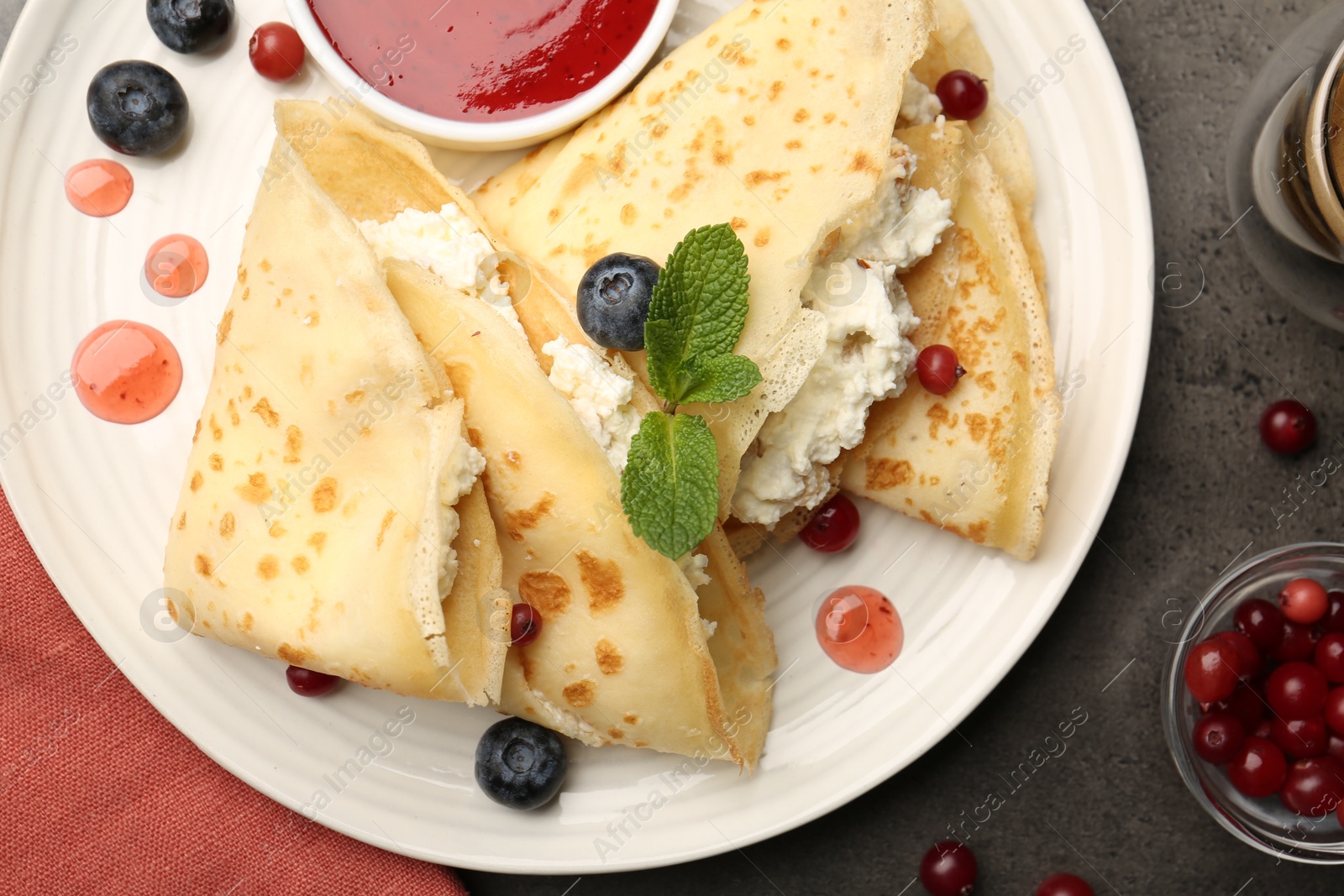 Photo of Delicious crepes with cottage cheese, jam, redcurrants and blueberries on grey table, flat lay