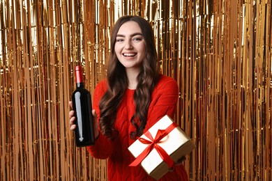 Photo of Happy young woman with gift box and bottle of wine against golden foil curtain