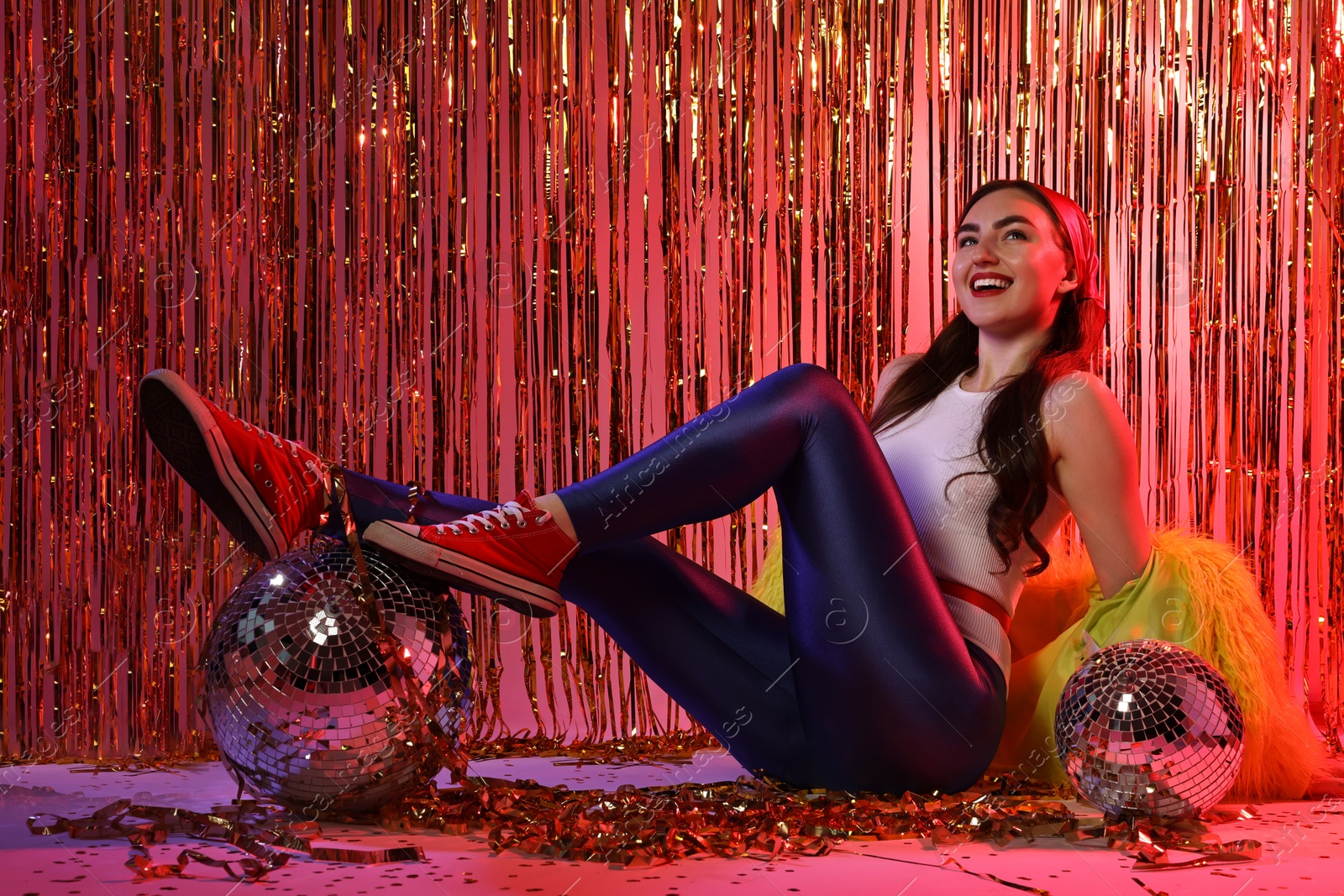 Photo of Beautiful young woman wearing retro outfit with tinsel and disco balls against golden foil curtain in neon lights