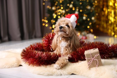 Photo of Cute dog in Santa hat with shiny tinsel and gift box on floor against blurred lights