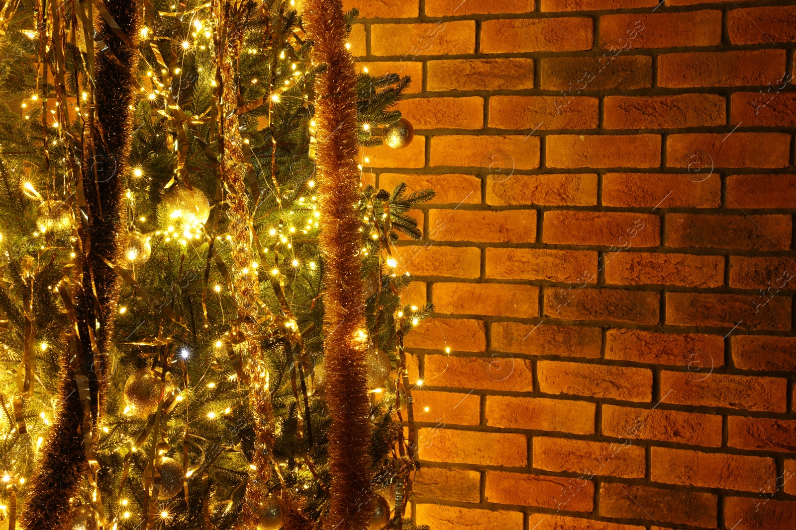 Photo of Shiny tinsels, baubles and festive lights on Christmas tree near brick wall. Space for text