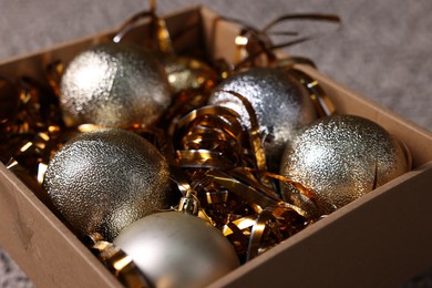 Photo of Shiny tinsels and baubles in cardboard box against blurred background, closeup