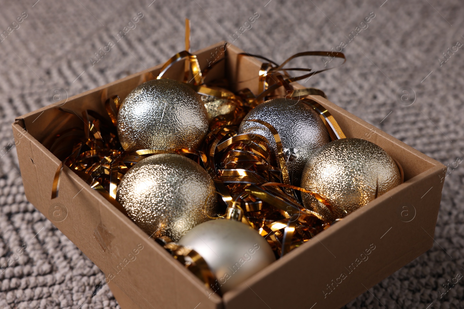 Photo of Shiny tinsels and baubles in cardboard box on carpet, closeup