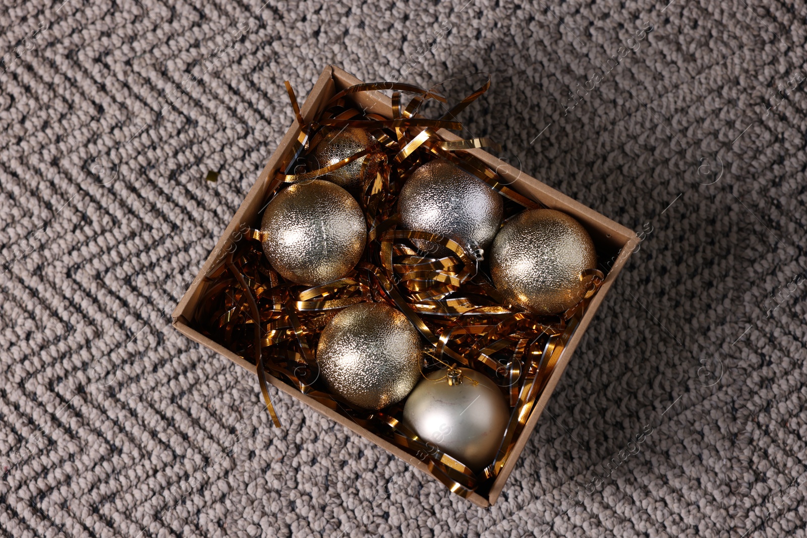 Photo of Shiny tinsels and baubles in cardboard box on carpet, top view