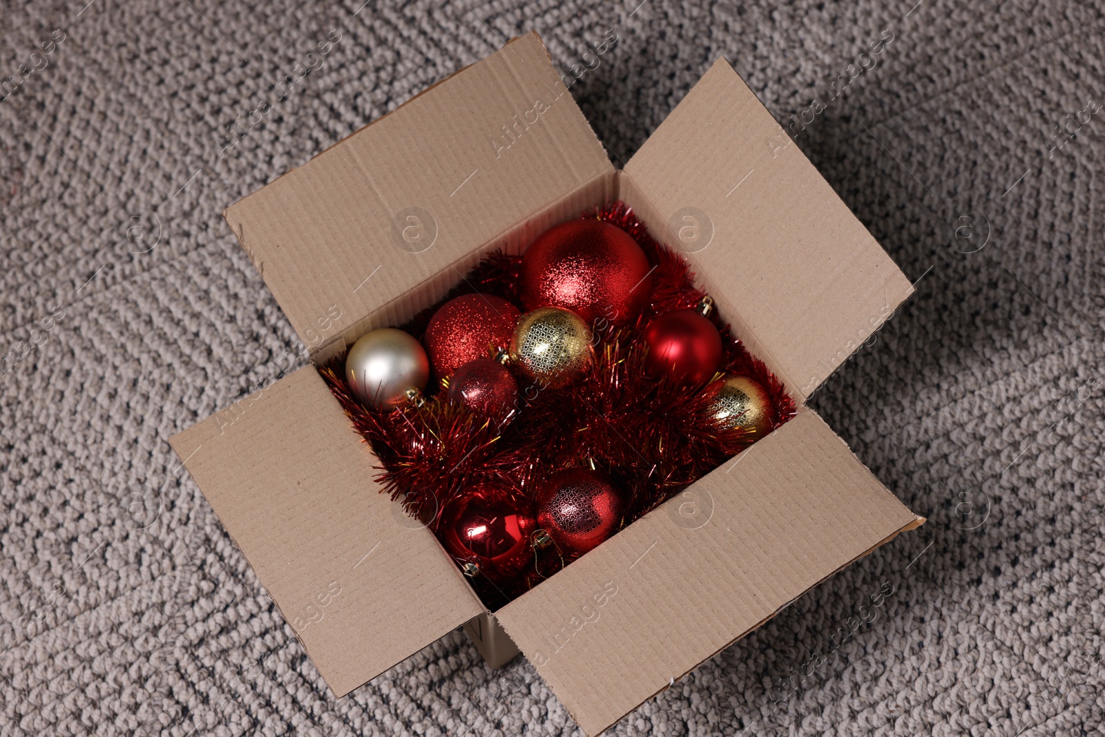 Photo of Shiny tinsels and baubles in cardboard box on carpet, above view