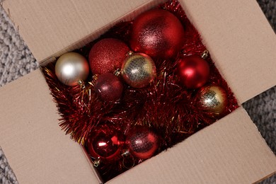 Photo of Shiny tinsels and baubles in cardboard box on carpet, top view